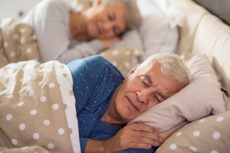 Casal idoso dormindo pacificamente na cama, simbolizando os benefícios de um sono profundo para a saúde, energia diária e equilíbrio mental.