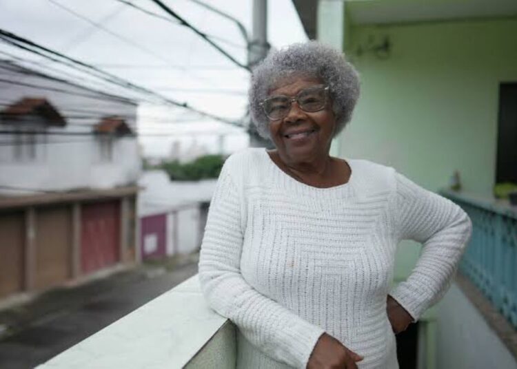 Mulher idosa sorrindo, com cabelos grisalhos e usando um suéter branco, apoiada na varanda, representando o tema de mulheres dominando a Previdência.