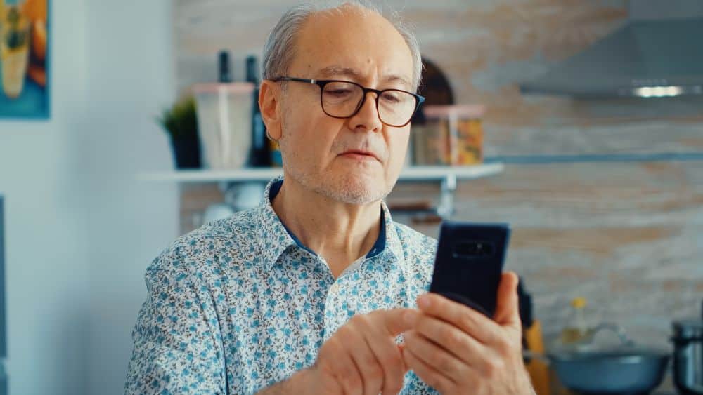 Homem idoso, em casa, usando um smartphone. A imagem representa a postagem de frases curtas de fim de semana no status do WhatsApp ou redes sociais.