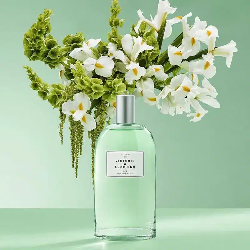 Frasco verde-claro do perfume Iris Nº3 LUMINOSO da Victorio & Lucchino em frente a um buquê de flores de íris brancas, representando o aroma intenso e refinado.