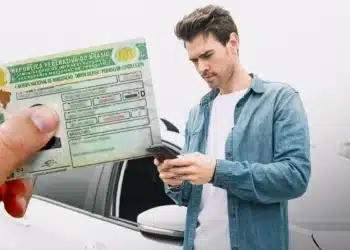 Motorista jovem ao lado de um carro verificando informações no celular, com a nova Carteira Nacional de Habilitação (CNH) física em destaque, representando a renovação do documento.