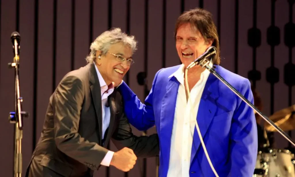 Caetano Veloso e Roberto Carlos se cumprimentando e sorrindo durante uma apresentação ao vivo, refletindo sua amizade e respeito mútuo.