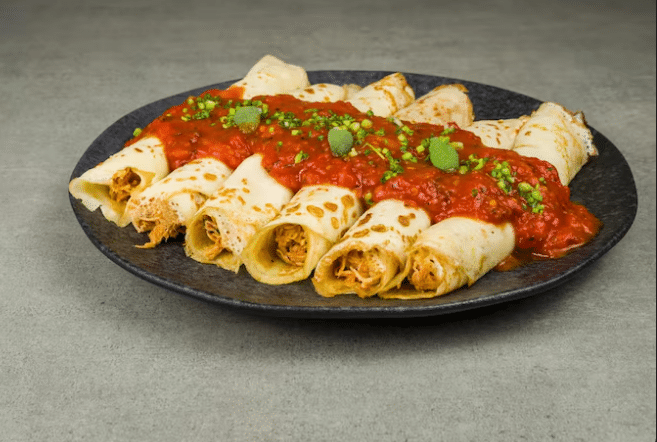 Seis panquecas enroladas e recheadas dispostas em um prato escuro, cobertas com molho de tomate vermelho vibrante e finalizadas com ervas frescas picadas.