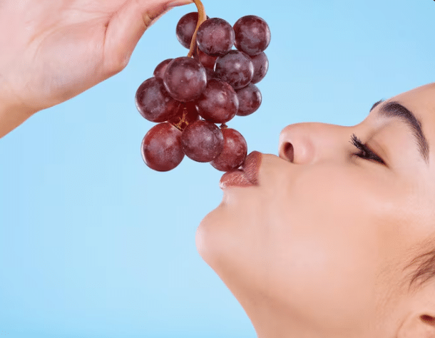 Perfil de uma mulher com olhos fechados aproximando os lábios de um cacho de uvas roxas segurado por uma mão, contra um fundo azul claro.