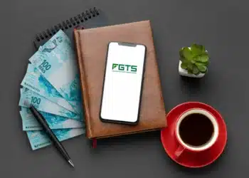 Smartphone mostrando logo do FGTS sobre caderno, dinheiro, caneta, xícara de café e planta