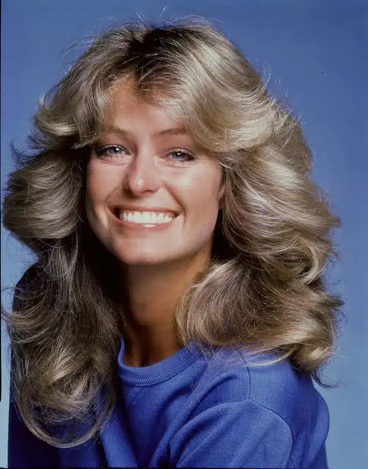 Farrah Fawcett com cabelo em camadas, franja mais curta no centro e pontas modeladas para fora, visual icônico dos anos 70.