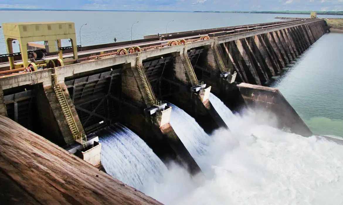 Barragem de concreto liberando grande volume de água com estrutura robusta e céu azul ao fundo