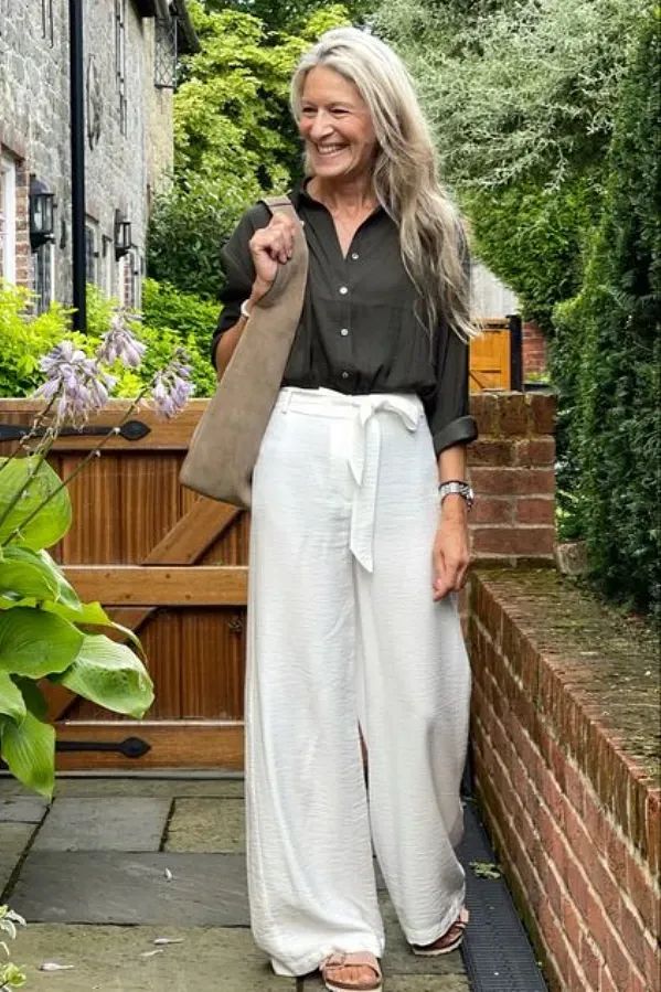 Mulher sênior sorridente com óculos de sol, vestindo uma saia midi estruturada (estilo pantacourt) em tons de verde oliva e branco, segurando uma bolsa de palha estruturada.