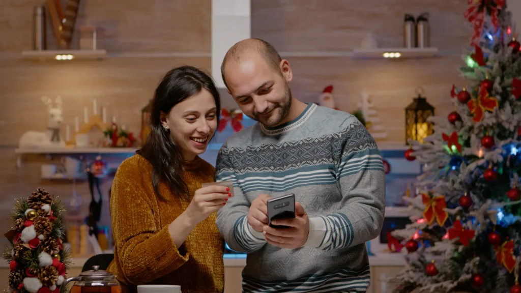 Casal sorrindo e compartilhando um momento de alegria, enquanto visualizam algo em um smartphone, com decoração de Natal ao fundo.