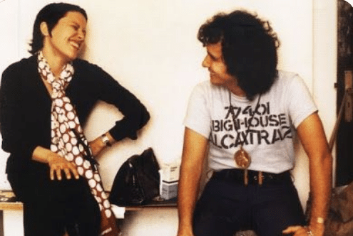 Duas pessoas conversando sorrindo sentadas, uma com lenço branco e preta, outra com camiseta branca e colar