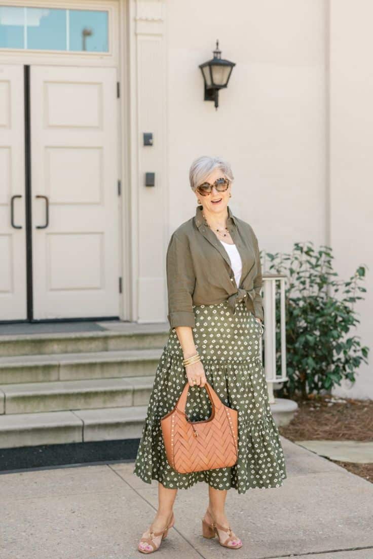 Mulher sênior elegante usando saia midi verde com estampa geométrica, camisa de linho amarrada na cintura e bolsa de couro, em frente a uma entrada clássica.