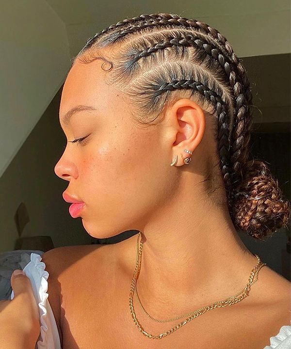 Mulher jovem com tranças nagô finas desenhadas na lateral e o cabelo preso em um coque baixo na nuca. O penteado é complementado por brincos e colares dourados que dão um toque de luz festivo.