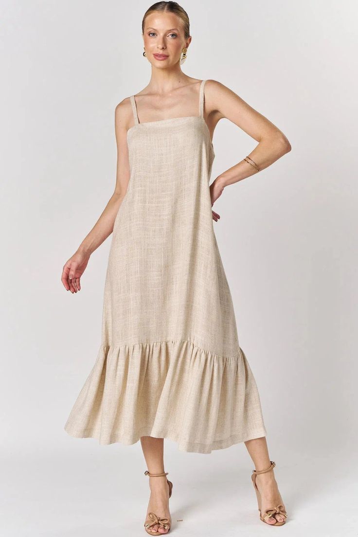 Modelo feminina vestindo um vestido midi de linho na cor bege areia, com alças finas e detalhe de babado na barra, posando contra um fundo branco.