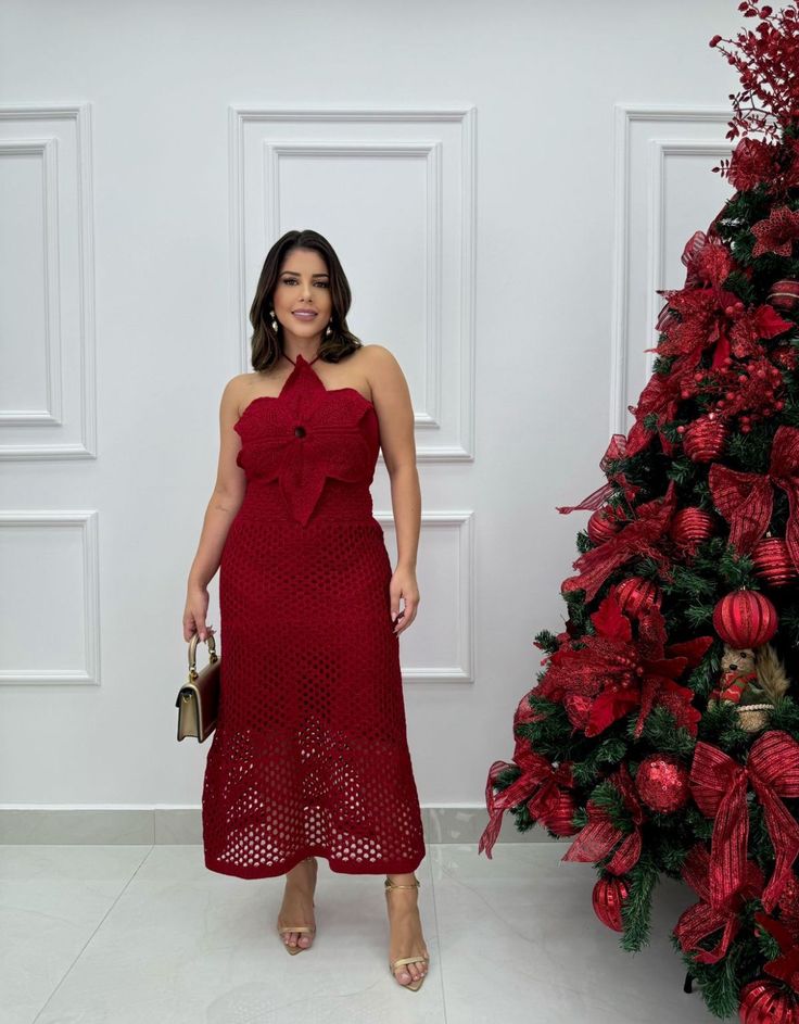 Mulher vestindo um elegante vestido vermelho de comprimento midi, com corte de alças largas e caimento sofisticado, em um cenário de decoração natalina festiva. O look é complementado por sandálias metalizadas e uma clutch.
