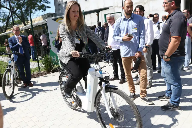Mulher pedalando bicicleta elétrica enquanto grupo de pessoas observa durante evento ao ar livre