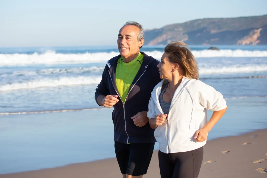 Casal de idosos caminhando na praia durante o verão, praticando exercício físico ao ar livre.