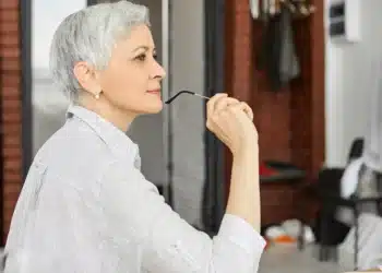 Mulher madura com cabelo curto grisalho em ambiente moderno, olhando pensativa enquanto segura seus óculos.