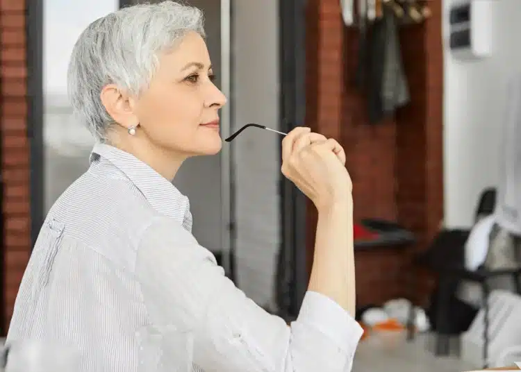 Mulher madura com cabelo curto grisalho em ambiente moderno, olhando pensativa enquanto segura seus óculos.