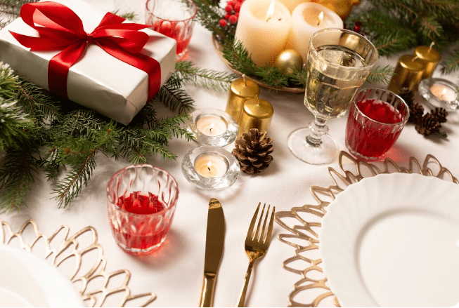 Mesa de Natal com prato branco, talheres dourados, velas acesas, pinha, copos com bebidas vermelhas e presente com laço vermelho