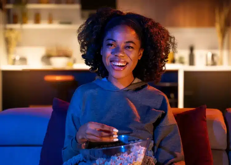 Mulher sorrindo enquanto assiste TV, segurando uma tigela de pipoca em ambiente aconchegante, com iluminação suave, simbolizando uma noite tranquila para maratonar doramas.