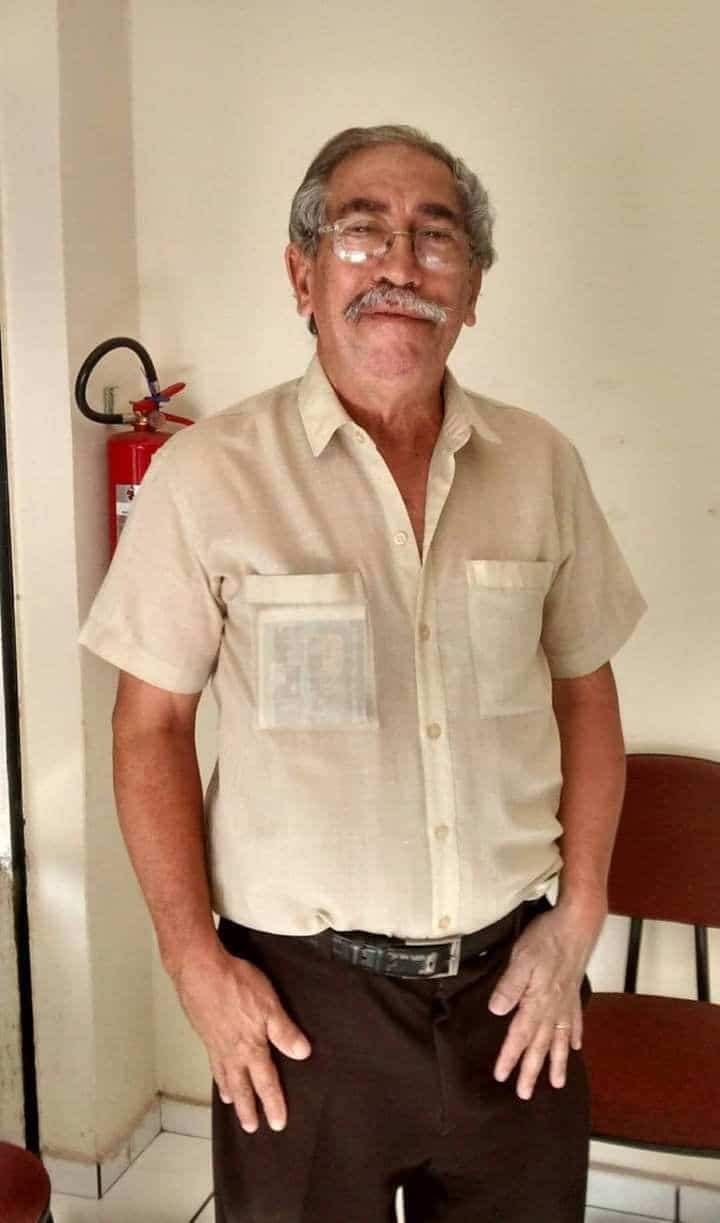 Homem idoso de óculos e bigode vestindo uma camisa de manga curta bege em tecido leve, com bolsos frontais, combinada com calça social escura.
