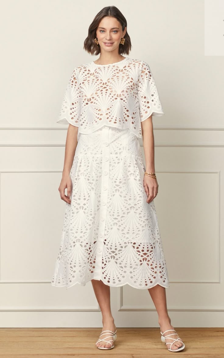 Modelo feminina vestindo um conjunto de duas peças em renda de algodão branca com padrões vazados em formato de leque, composto por blusa de mangas curtas e saia midi com botões frontais.
