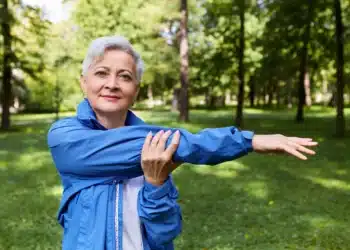 Idosa praticando exercícios ao ar livre, em um ambiente natural, usando um casaco azul, realizando alongamento de braço.