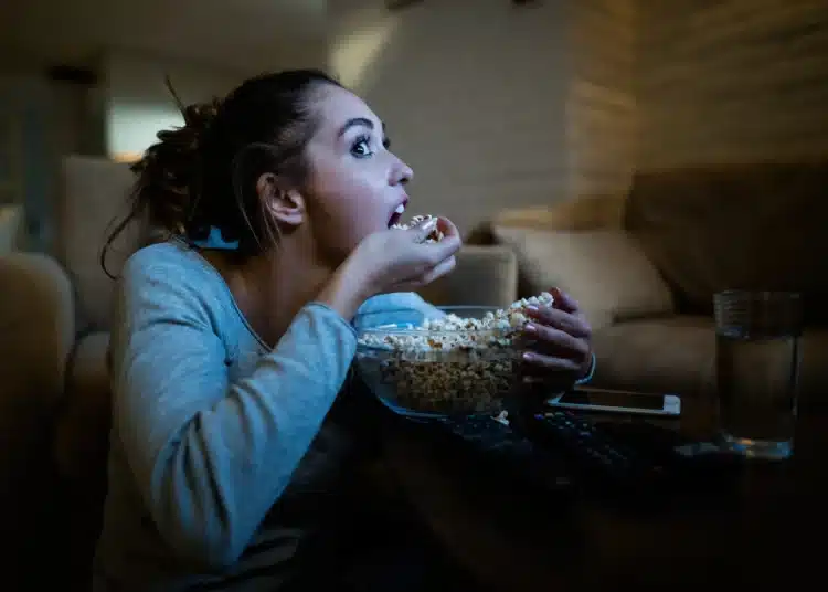 Mulher assistindo a um filme comendo pipoca, em um ambiente aconchegante, com foco no entusiasmo de quem está assistindo