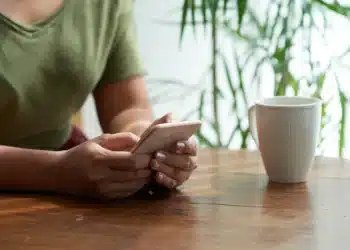 Pessoa segurando um smartphone com uma xícara de café sobre a mesa, rodeada por plantas verdes em um ambiente acolhedor.