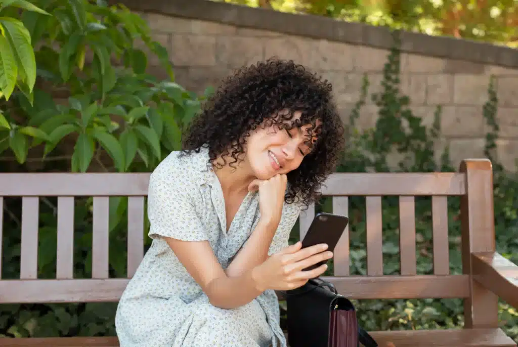 Mulher sorrindo sentada em um banco ao ar livre lendo mensagens inspiradoras no celular.