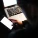 Pessoa usando celular à noite com laptop ao fundo, simbolizando o risco de fraudes online durante as festas de fim de ano.