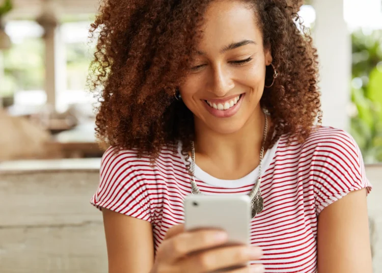 Pessoa sorrindo enquanto lê mensagem no celular, representando um momento carinhoso de bom dia.