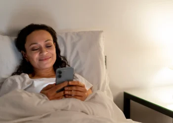 Mulher sorrindo na cama enquanto envia uma mensagem no celular, preparando-se para descansar após um dia cansativo de segunda-feira.