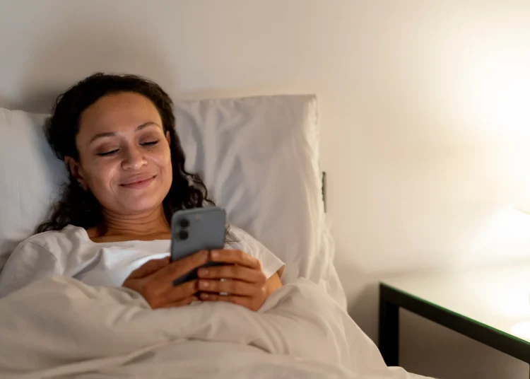 Mulher sorrindo na cama enquanto envia uma mensagem no celular, preparando-se para descansar após um dia cansativo de segunda-feira.