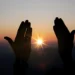 Silhueta de mãos elevadas em frente ao pôr do sol, transmitindo um momento de conexão espiritual e fé.