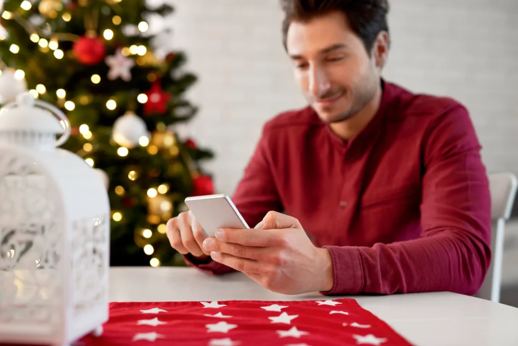 Homem usando smartphone em ambiente natalino com árvores de Natal e luzes brilhantes, perfeito para mensagens de fim de ano.