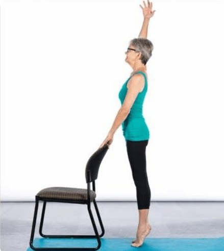 Idosa fazendo exercício de alongamento em pé segurando cadeira e elevando braço