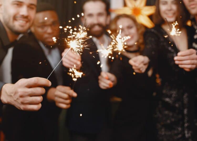 Grupo de amigos multiculturais vestidos com roupas de festa escuras, sorrindo e segurando velas faíscas acesas (sparklers) em um ambiente festivo com luzes douradas ao fundo.