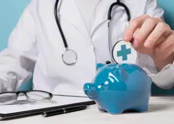 Médico segurando moeda simbólica de saúde enquanto coloca em um cofrinho azul, representando economia e contestação de reajustes de plano de saúde.