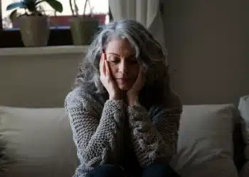 Mulher madura sentada no sofá, com expressão de cansaço e sobrecarga emocional, representando o esquecimento de si ao cuidar da família.