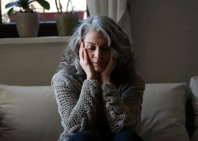 Mulher madura sentada no sofá, com expressão de cansaço e sobrecarga emocional, representando o esquecimento de si ao cuidar da família.