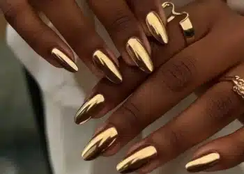 Unhas com esmalte cromado dourado e anéis dourados em destaque.