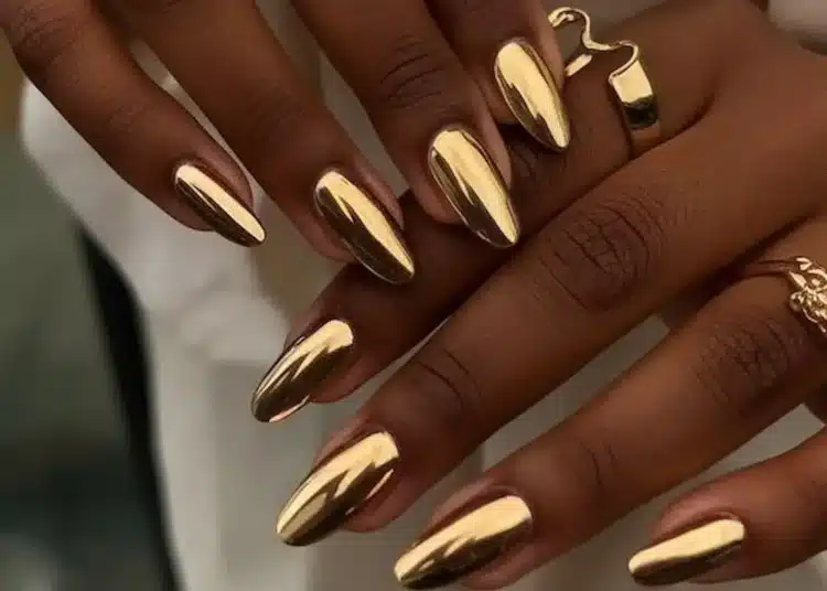 Unhas com esmalte cromado dourado e anéis dourados em destaque.