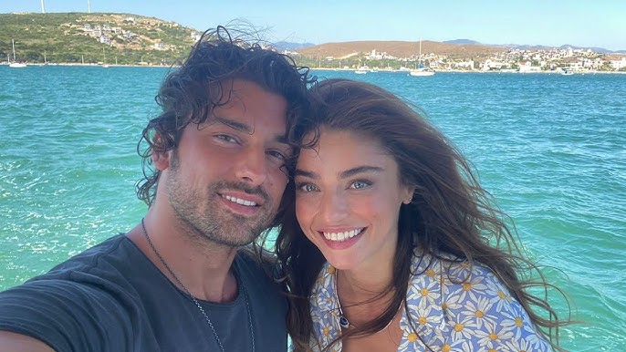 Selfie sorridente dos atores turcos Ayça Ayşin Turan e Alp Navruz a bordo de um barco, com o mar azul-turquesa e uma costa com casas brancas e barcos ao fundo. Ayça veste uma camisa estampada com margaridas.