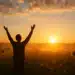 Pessoa de braços erguidos em um campo ao nascer do sol.