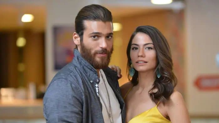 O casal de atores turcos Can Yaman e Demet Özdemir posando juntos. Can tem barba, cabelos longos presos e veste uma camisa cinza. Demet está ao seu lado com cabelos castanhos ondulados, usando um vestido amarelo e brincos de folha verde.