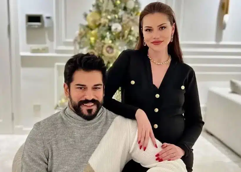 fahriye evcen burak ozcivit casamento novela calikusu O casal de atores turcos Burak Özçivit e Fahriye Evcen em um ambiente interno decorado. Burak está sentado, vestindo um suéter de gola alta cinza e branco, e Fahriye está em pé atrás dele, com a mão em seu ombro, usando um cardigã preto com botões dourados e um colar de corrente.