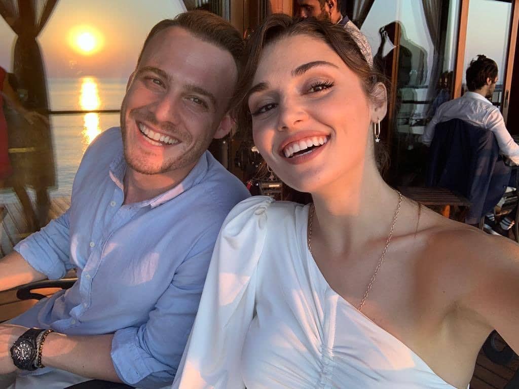 Selfie sorridente dos atores turcos Kerem Bürsin e Hande Erçel durante o pôr do sol. Kerem usa camisa azul clara e Hande veste uma blusa branca de um ombro só. Ao fundo, o reflexo do sol no mar.