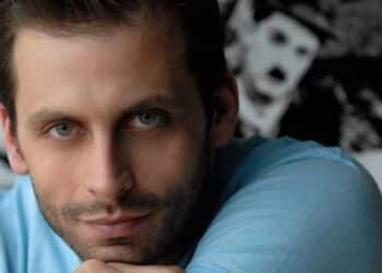 Close do rosto do ator Henri Castelli, com olhos azuis em destaque, barba rala e expressão serena. Ele está apoiando o queixo sobre a mão, vestindo uma camiseta azul clara.