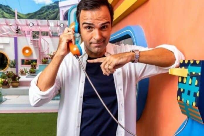 Homem atende telefone colorido no cenário do BBB, sorrindo e apontando para o aparelho
